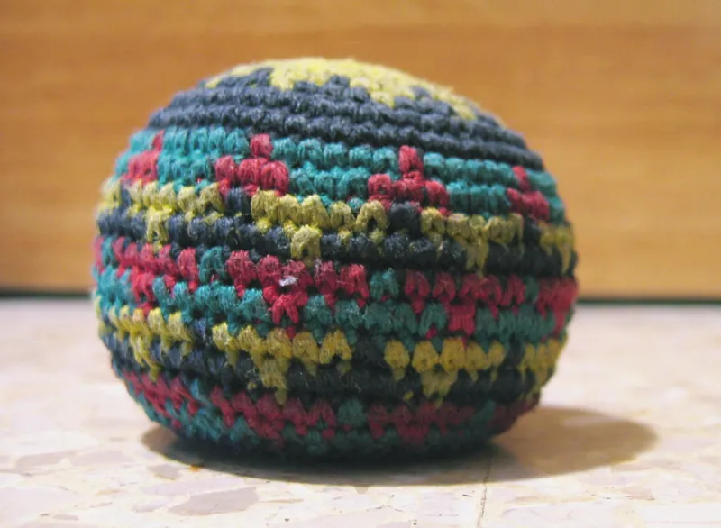 A crocheted footbag with red, green, yellow and dark blue stripes on a stone floor